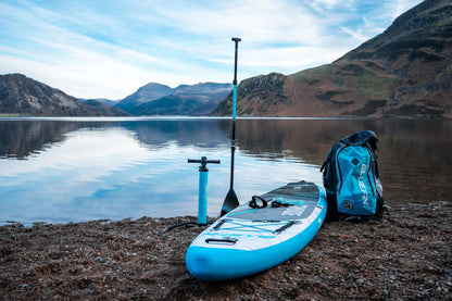 Inflatable paddleboard in blue with a textured deck pad, pump, paddle, and backpack by a lake.