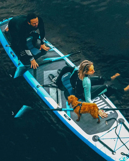 Two people on a blue inflatable paddleboard with a small dog, one person paddling and the other sitting.