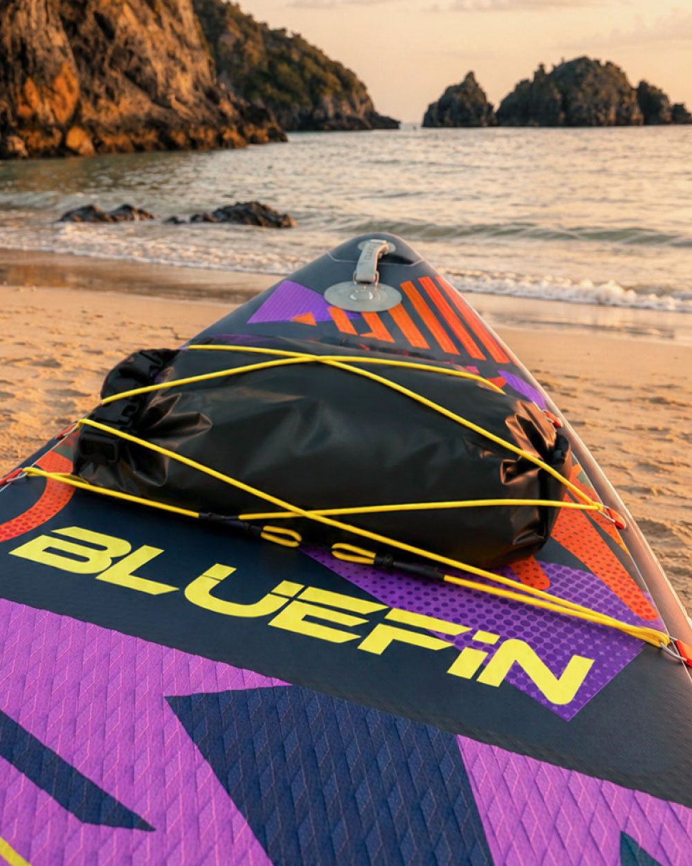 Inflatable paddleboard on beach with black dry bag, colorful design, and sunset in background