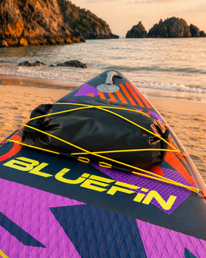 Inflatable paddleboard on beach with black dry bag, colorful design, and sunset in background