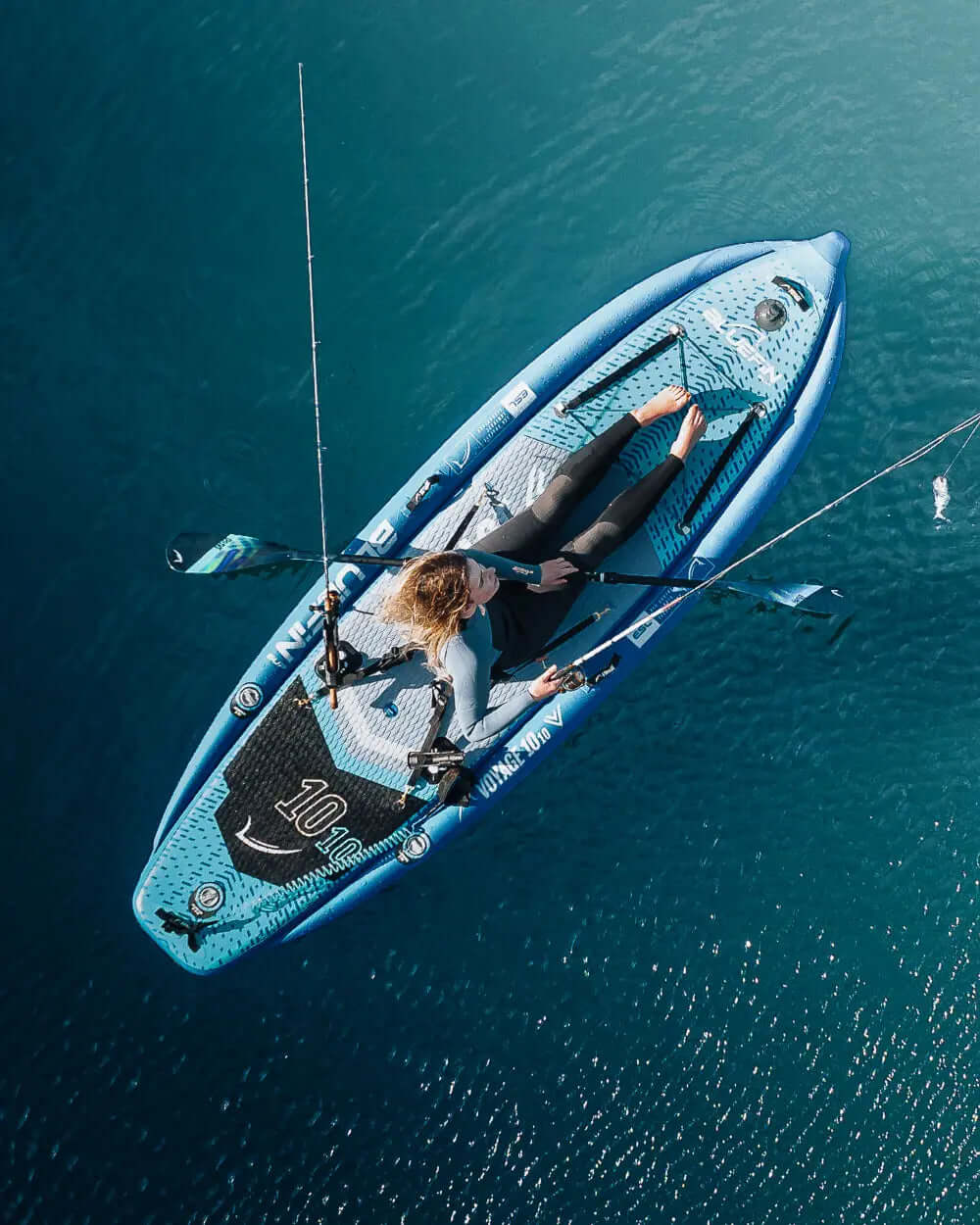 Inflatable paddleboard with a textured deck, fishing rod, and a person sitting on it in blue water.