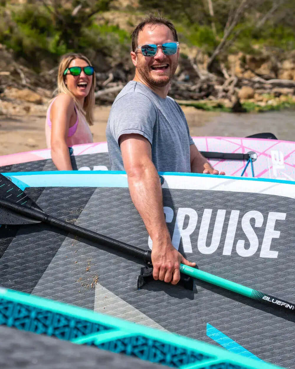 Two people smiling by inflatable paddleboards on a beach, one holding a paddle, sunny day.